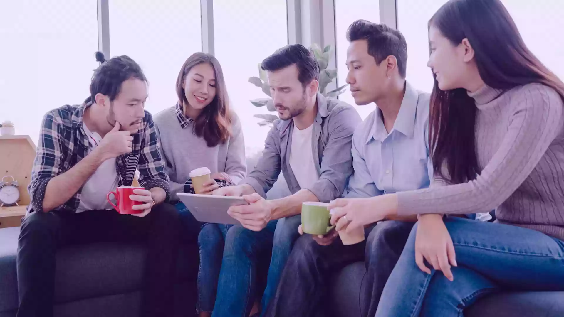 Five colleagues sitting together on a sofa, holding coffee cups and attentively viewing content on a tablet, representing collaborative planning and engagement with Cinema8’s interactive video hosting tools.