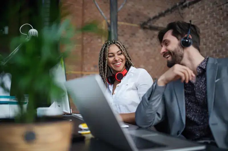 Two students smiling and collaborating at their laptops in a modern workspace, both wearing headsets. Captures the dynamic, remote-ready potential of video hosting for educational institutions, supporting teamwork on lecture content, internal updates, and secure faculty-led sessions.